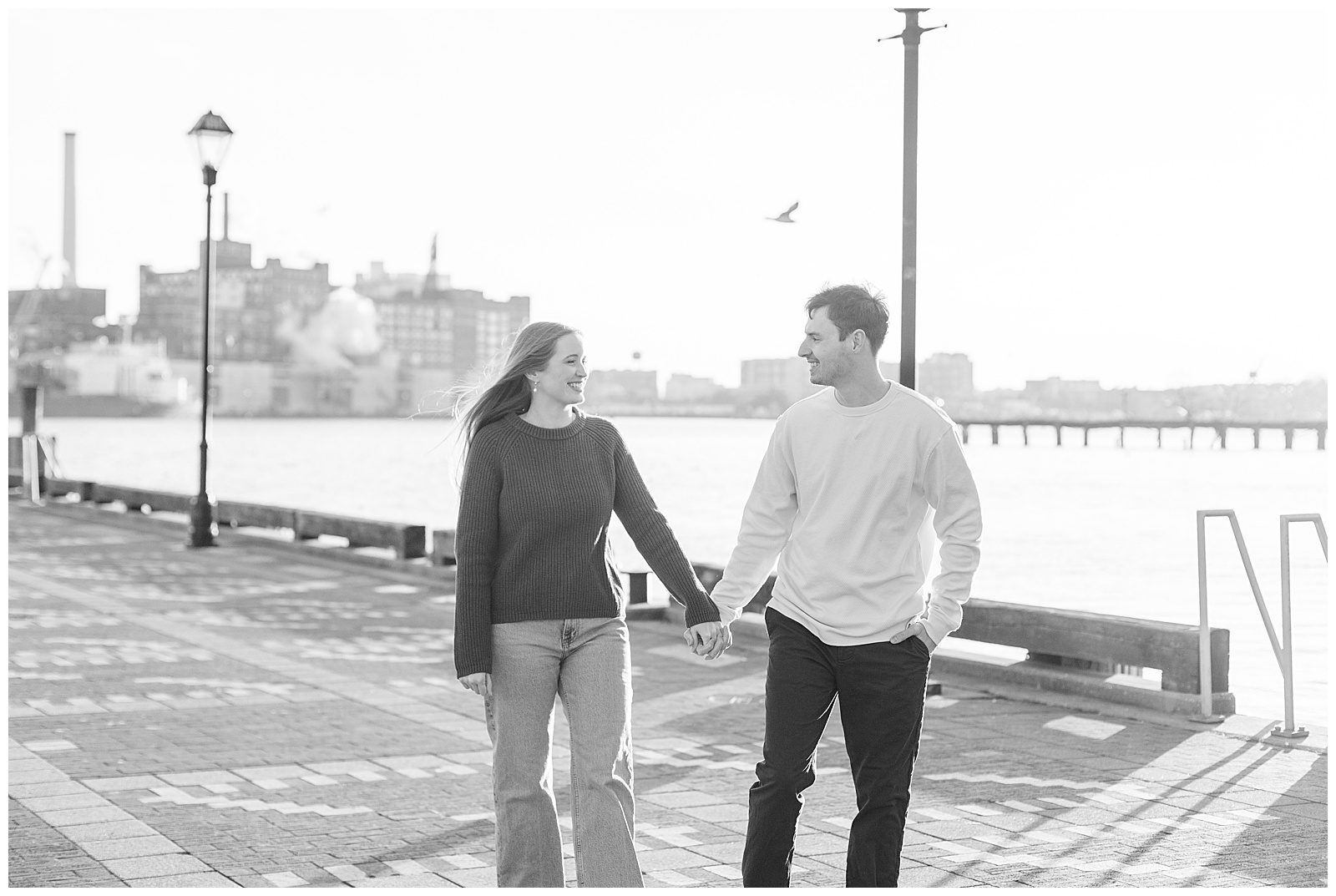 black and white photo of couple holding hands along walkway by chesapeake bay for this Fells Point Engagement Session