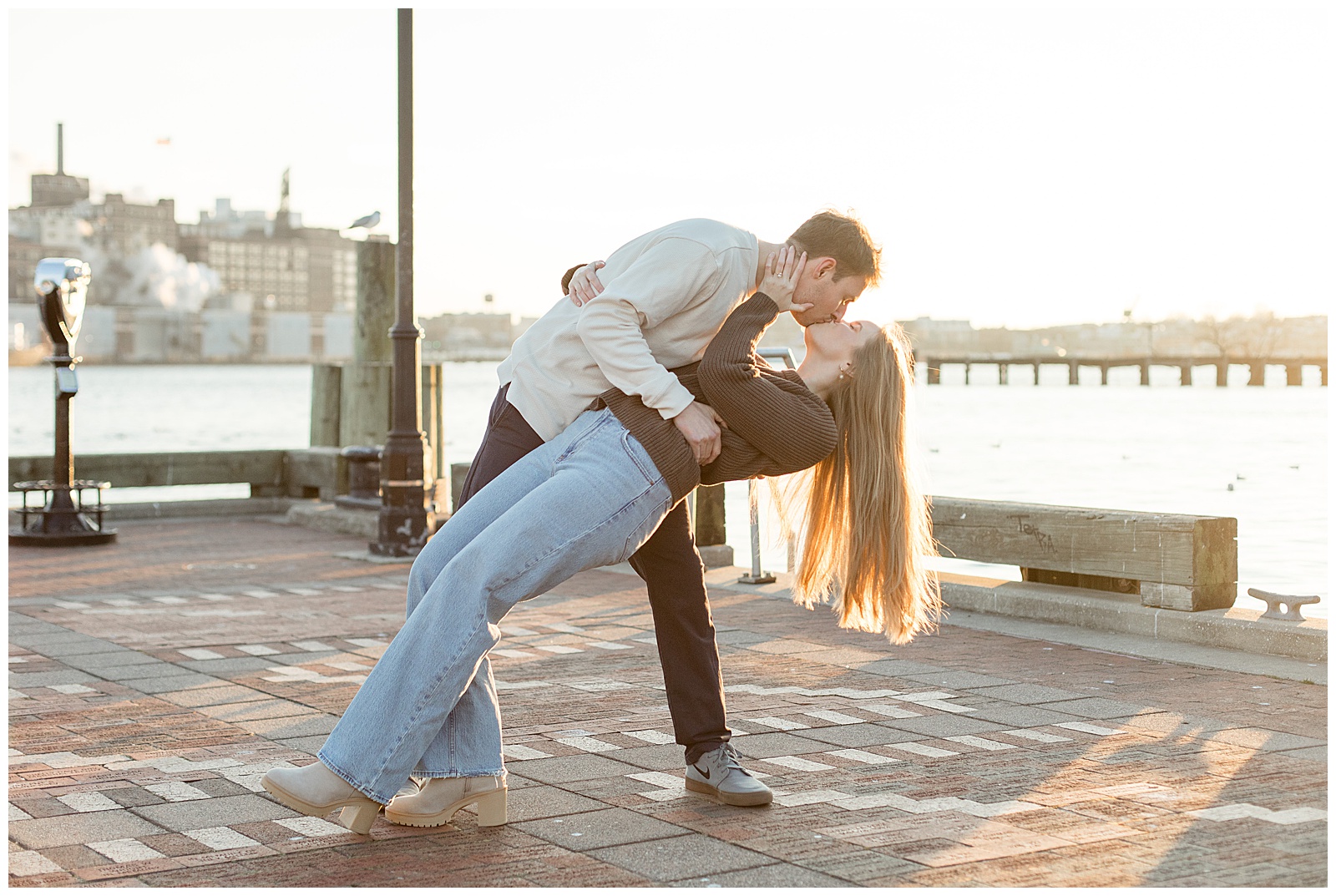 man dipping woman back as they kiss beside chesapeake bay at sunset for this Fells Point Engagement Session