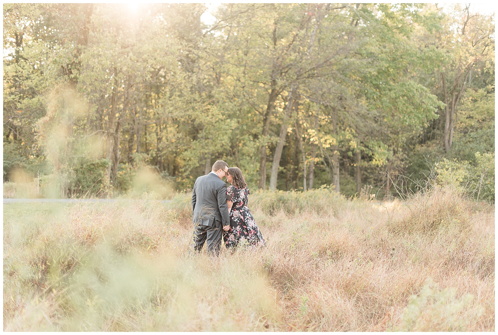 couple touching foreheads, standing in a field of grasses with light coming through the trees behind