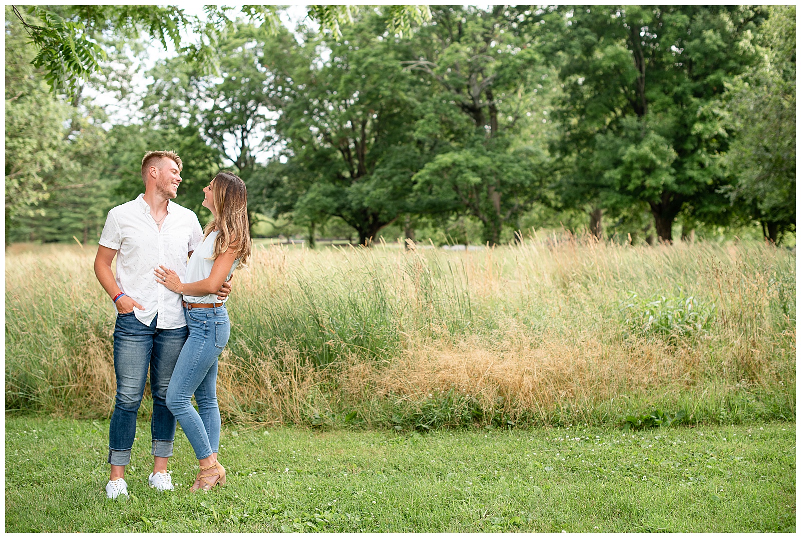 couple standing with arms around each other smiling at each other in front of tall grass field