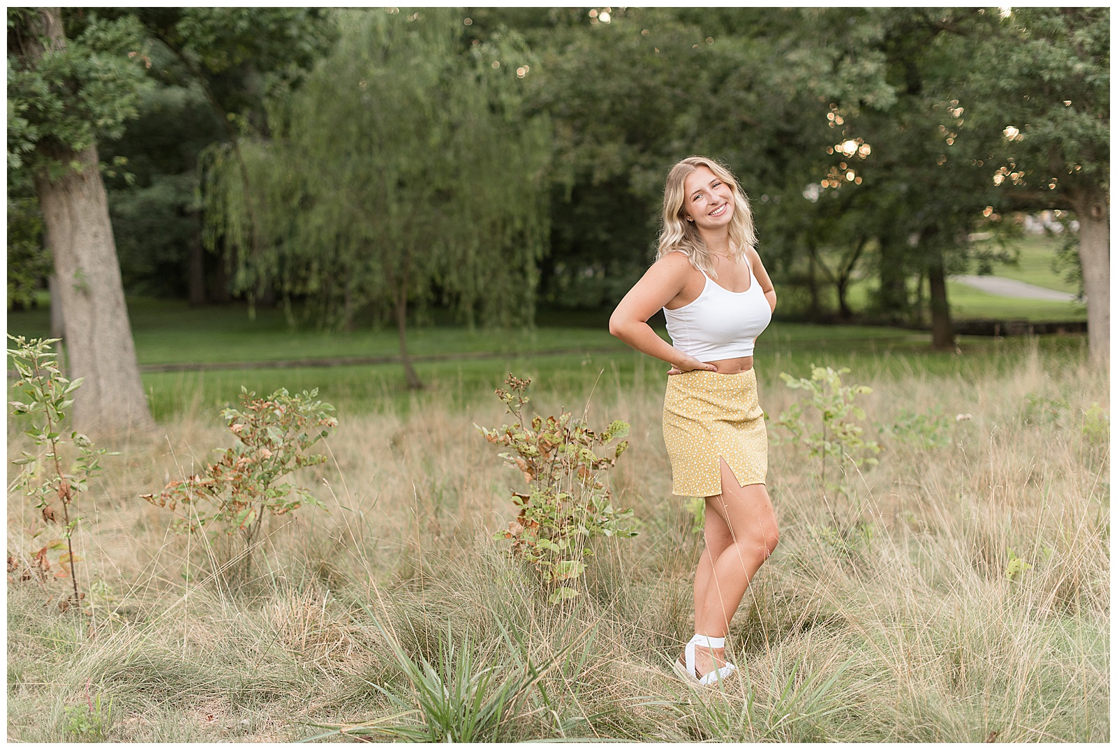 senior smiling with hands on hip while she stands in field of wild grasses