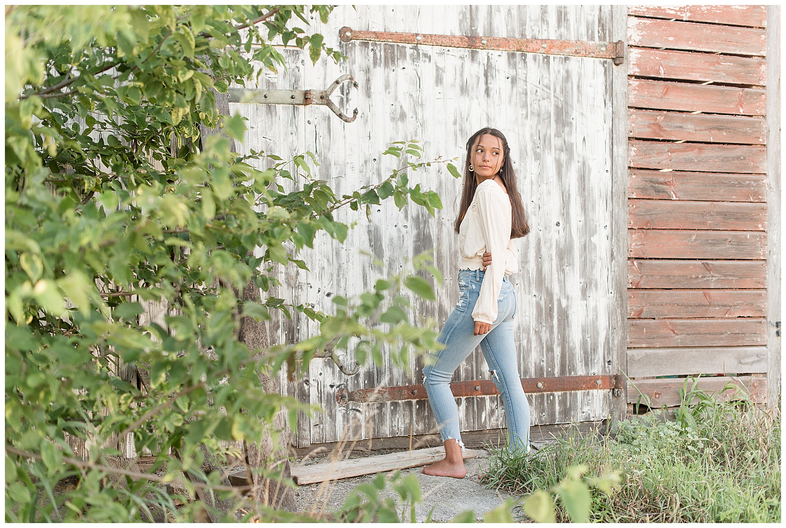 senior holding arm behind back and looking over shoulder while standing in front of old barn with white wood door