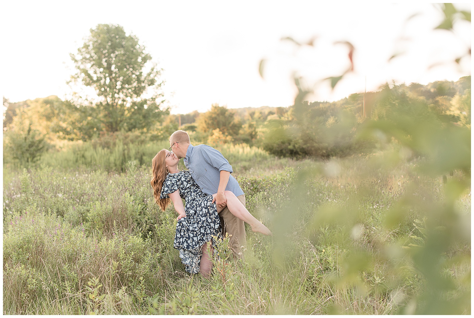 guy in blue shirt and Khakis dipping girl in navy blue floral dress surrounded by tall wild grasses