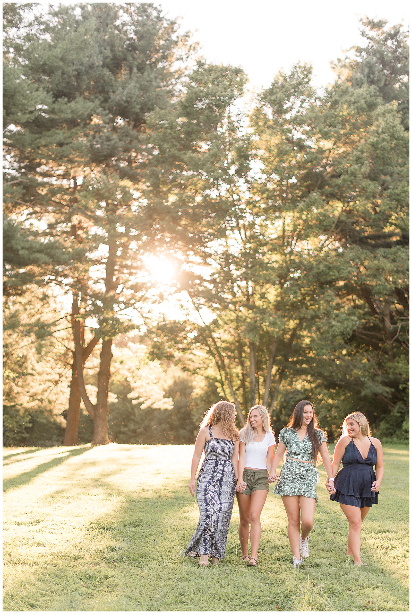 four senior girls linking arms walking towards camera in Lancaster County central park with sunlight shining through trees behind them