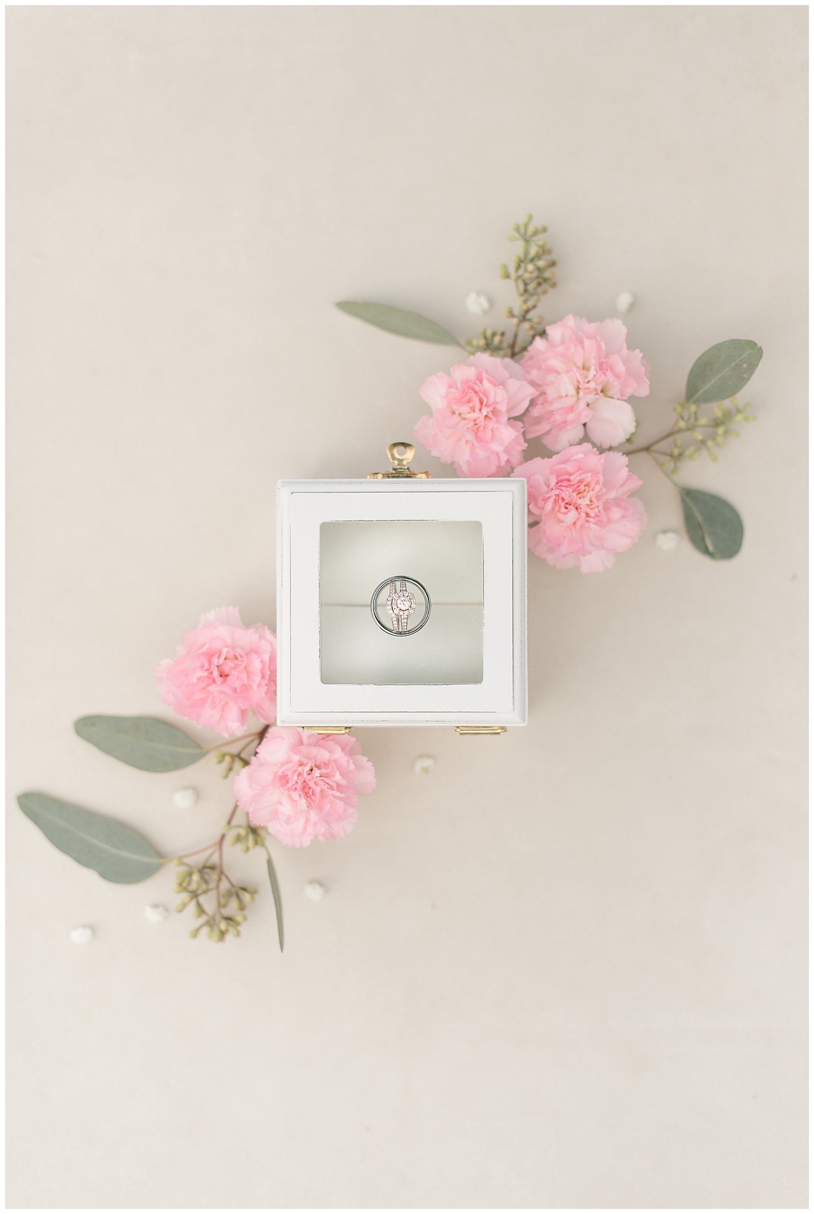 close up photo of bride's wedding ring inside small white box with clear lid and a small cluster of pastel pink carnations and eucalyptus at top right corner and bottom left corner of box at Lakefield Weddings in Manheim, PA