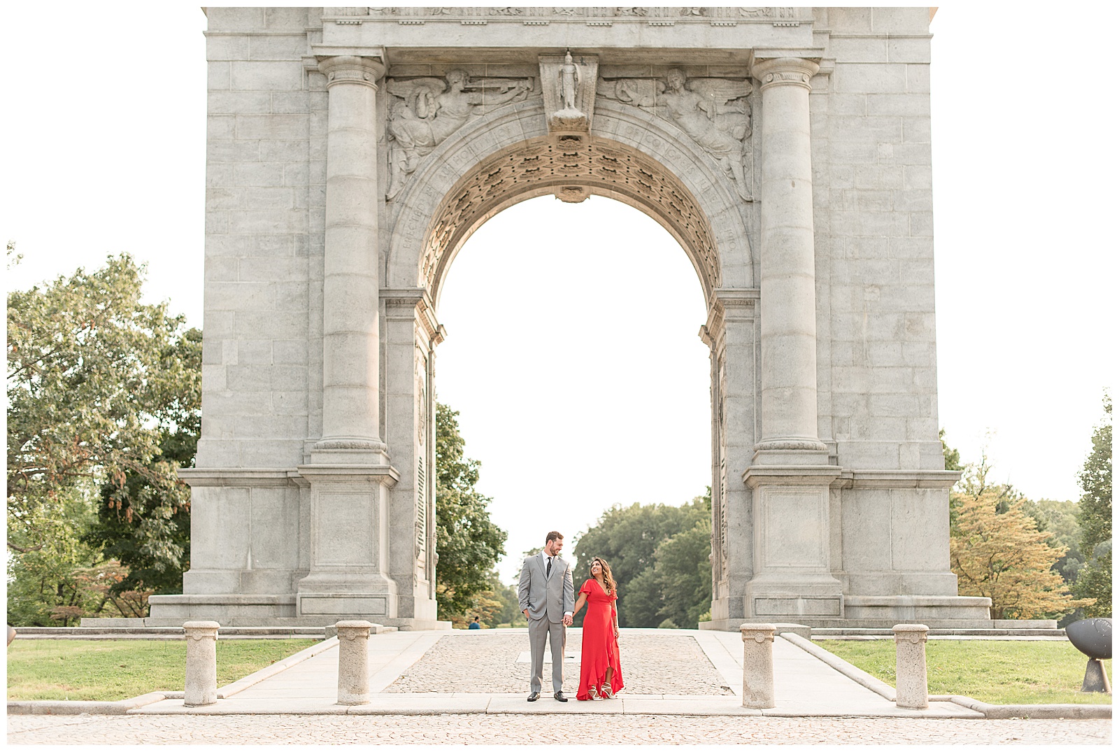 couple is standing underneath large beautiful archway with the guy on the left in a light gray suit and the girl on the right in a lovely red floor length dress and they are holding hands and looking at the camera at the National Memorial Arch in Valley Forge, PA