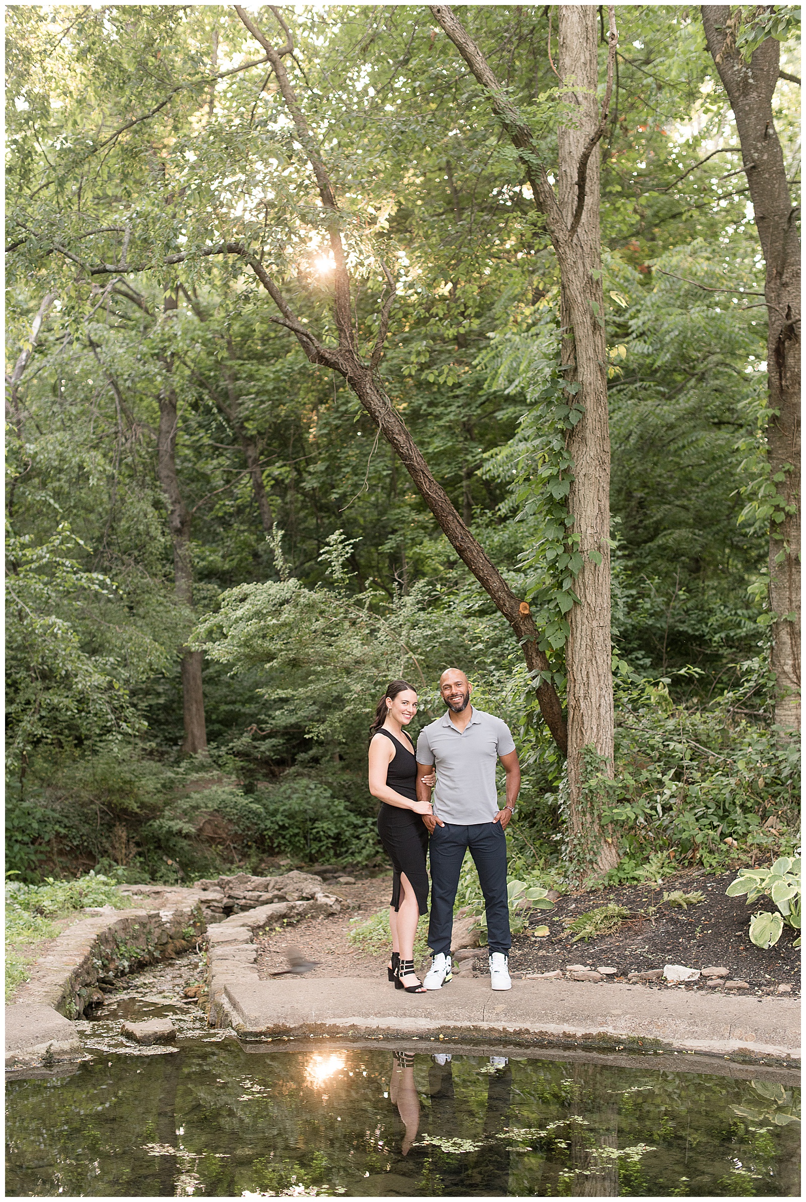 engaged couple standing by pond smiling at camera in wooded area at fonthill castle near philadelphia