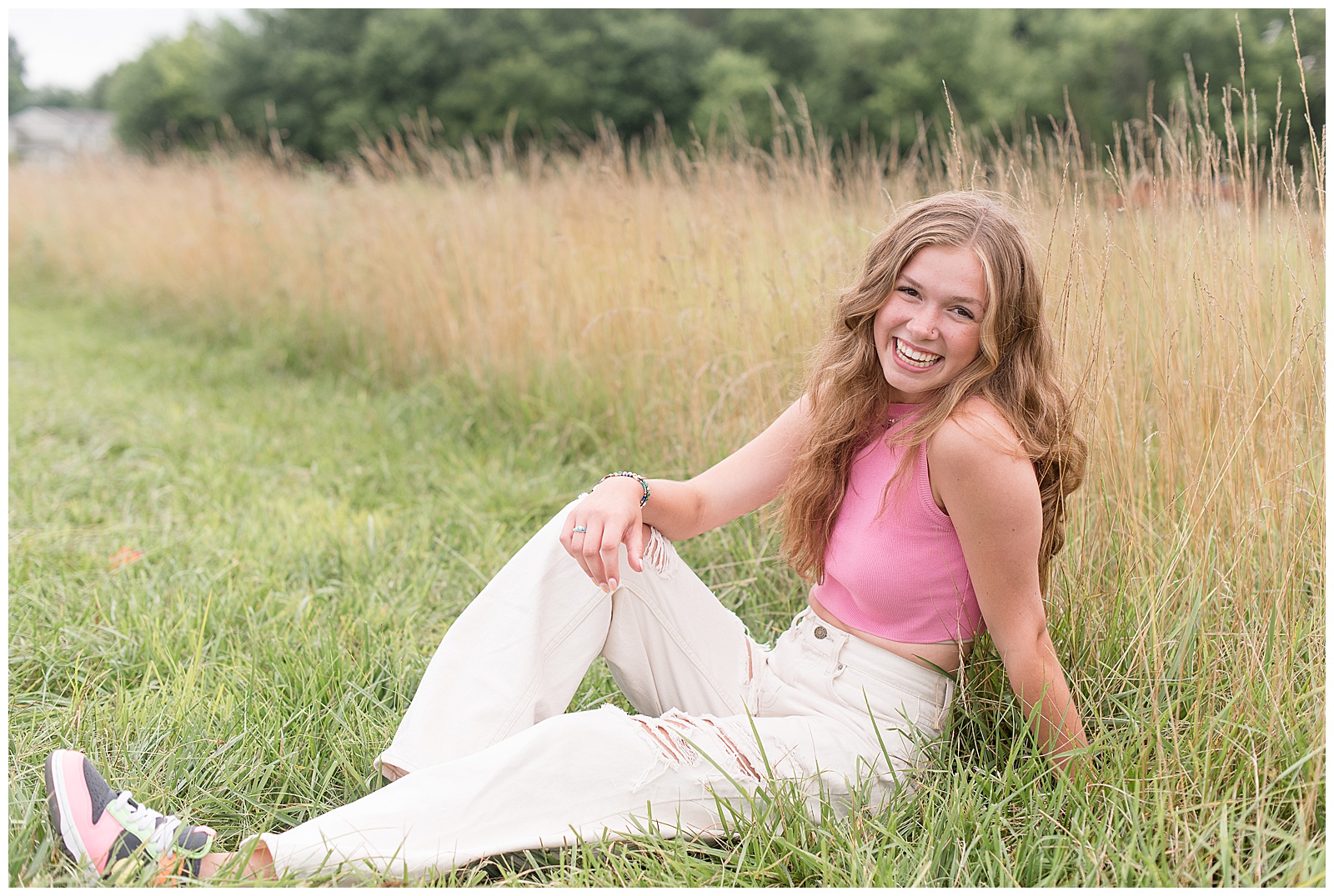 senior girl wearing pink top and white jeans sitting in grassy field with right arm resting on her bent right leg in lancaster pennsylvania