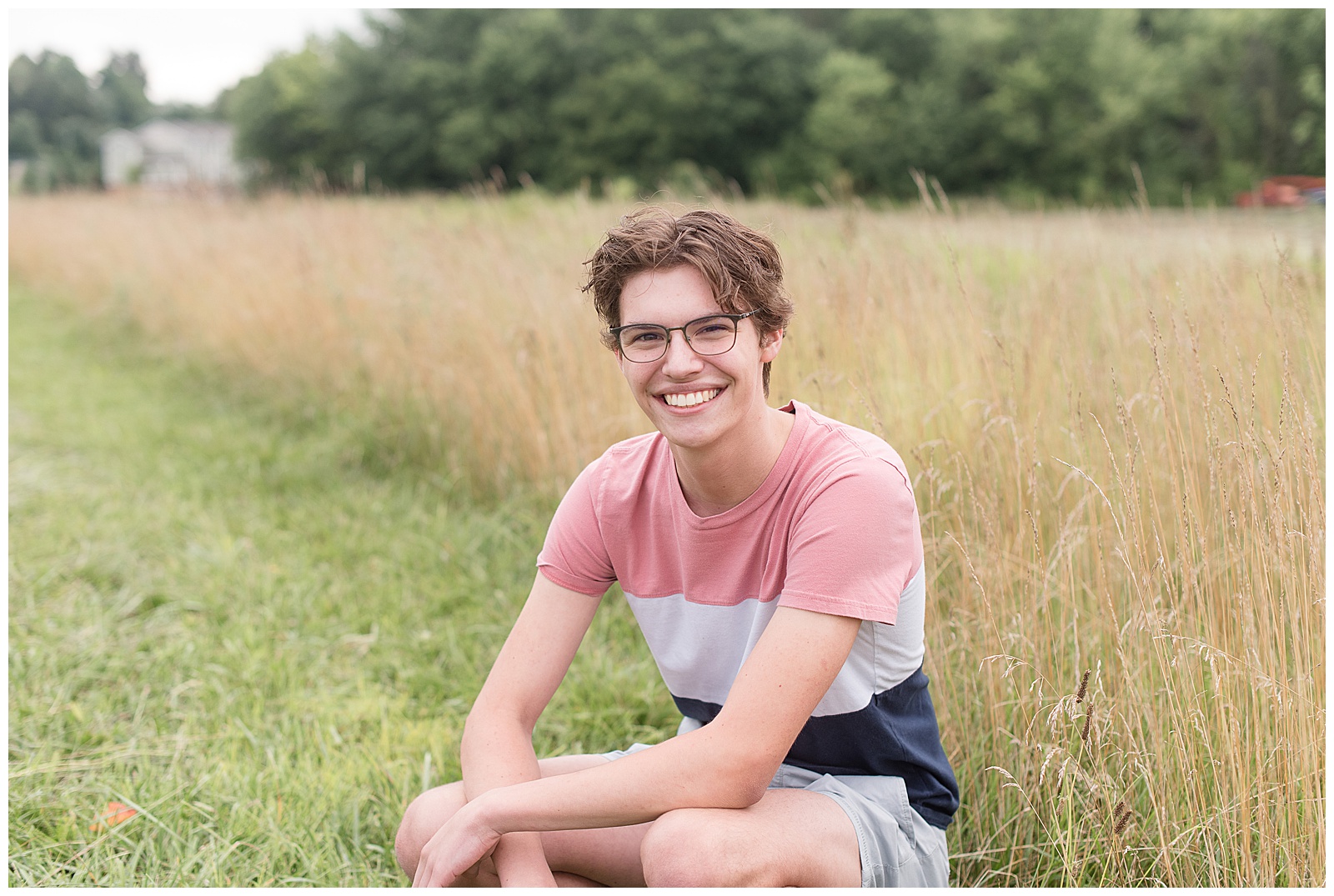 senior guy spokesmodel crouching down next to grassy field smiling wearing striped shirt and shorts