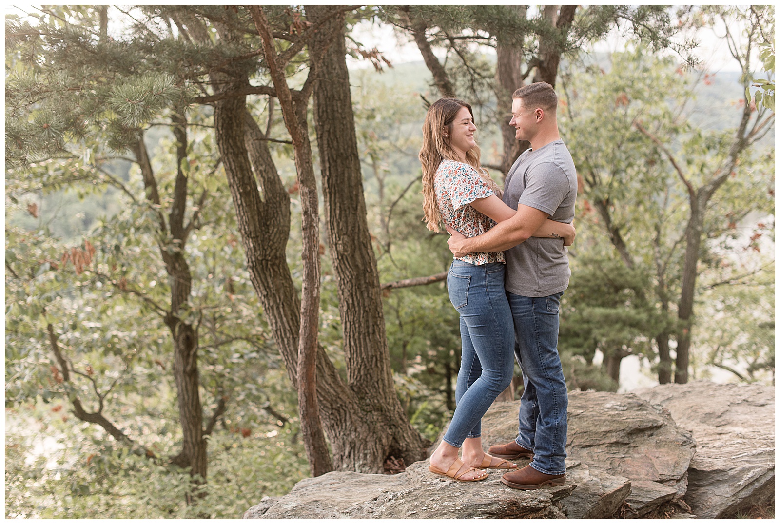 engaged couple hugging as they smile at each other standing on large rock by trees at pinnacle overlook 
