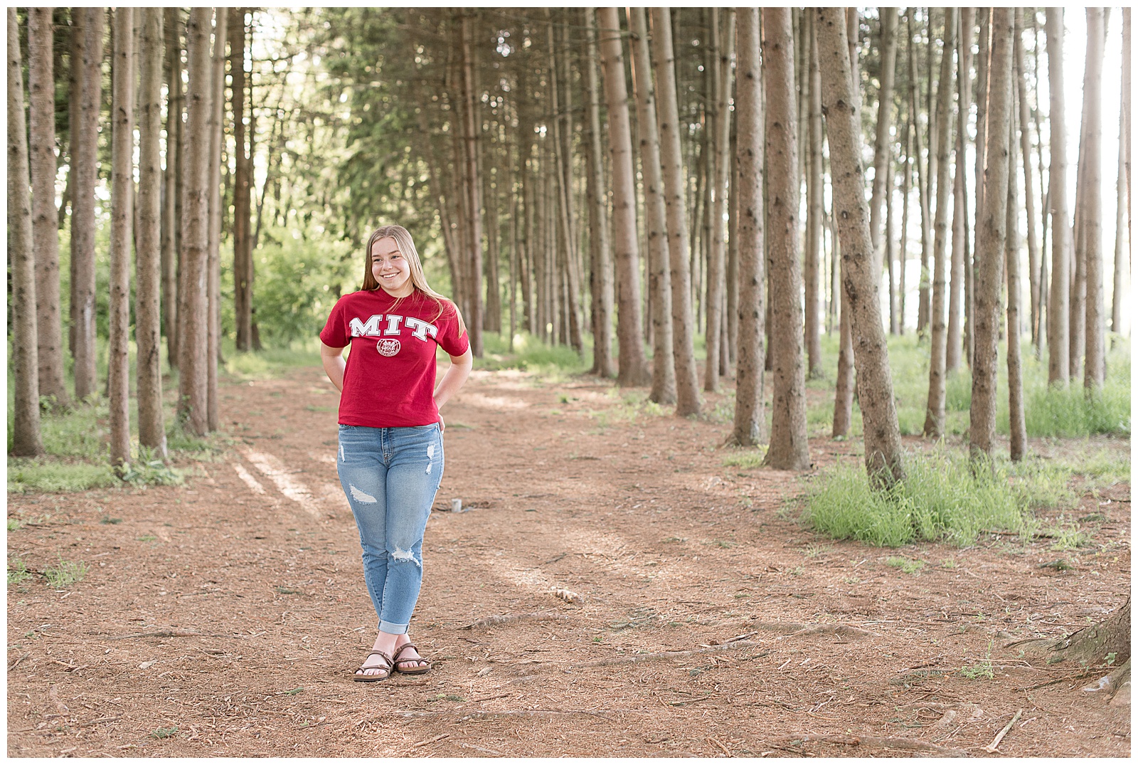 senior girl in red MIT tshirt and blue jeans standing among pine tree grove at overlook park