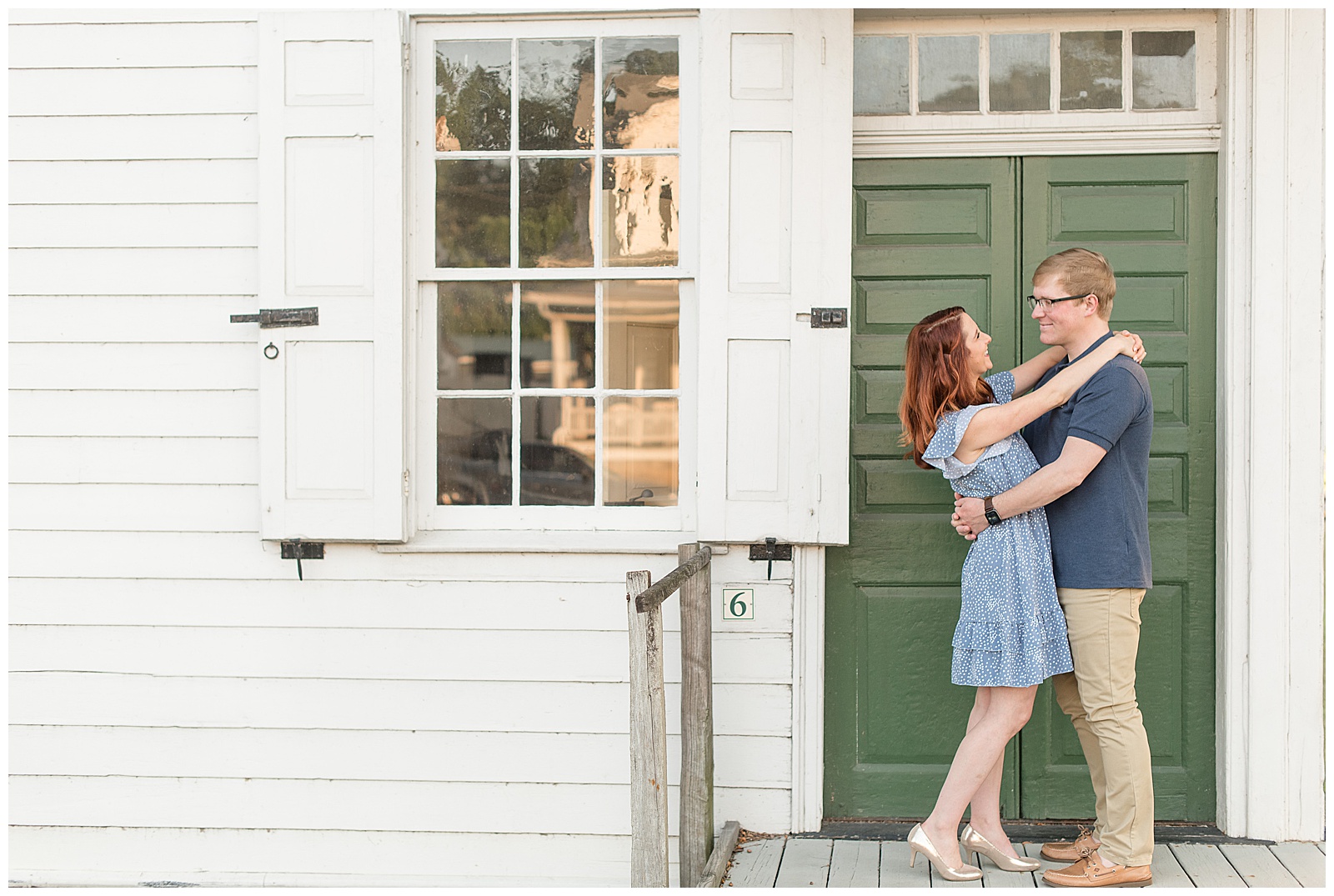 couple hugging and smiling as they stand on wooden porch smiling by white building with green door at ephrata cloister