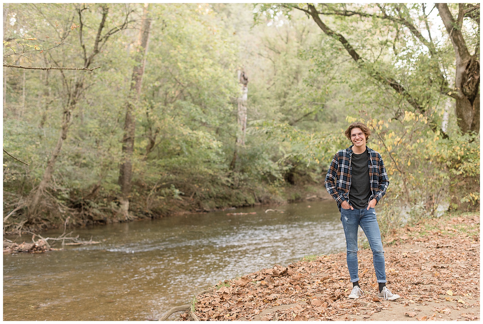 senior guy standing along conestoga river on dried leaves with hands in his pockets smiling at lancaster county park