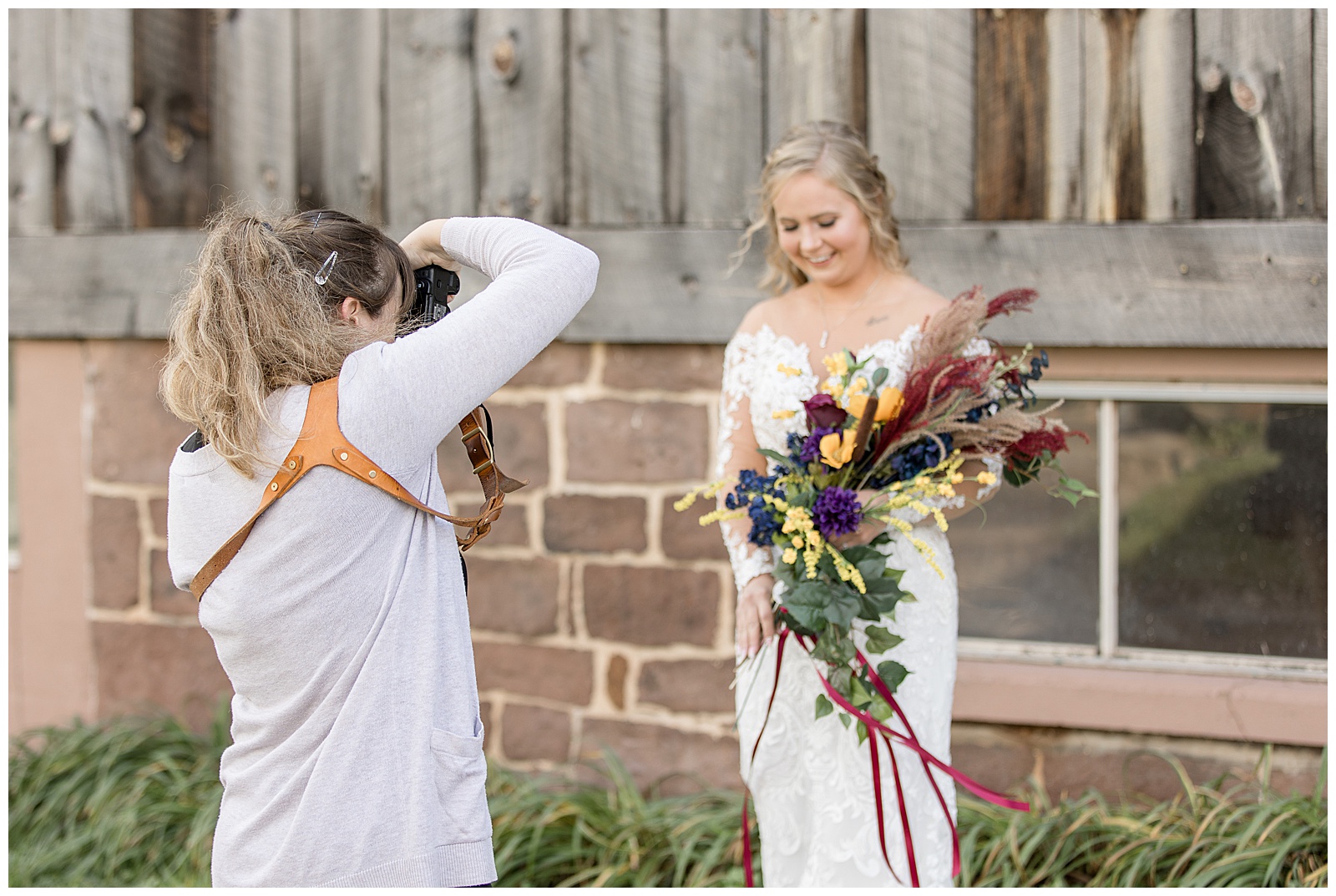 photographer capturing beautiful bride holding colorful fall bouquet by rustic wooden barn in york, pennsylvania