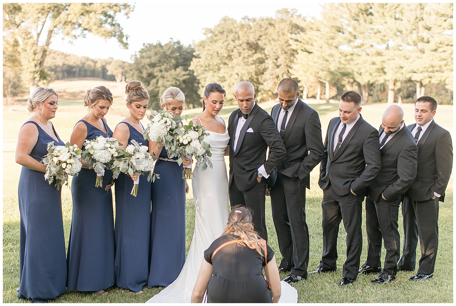 couple surrounded by bridal party as they all look down at photographer who's adjusting bride's dress train in front of them in new jersey