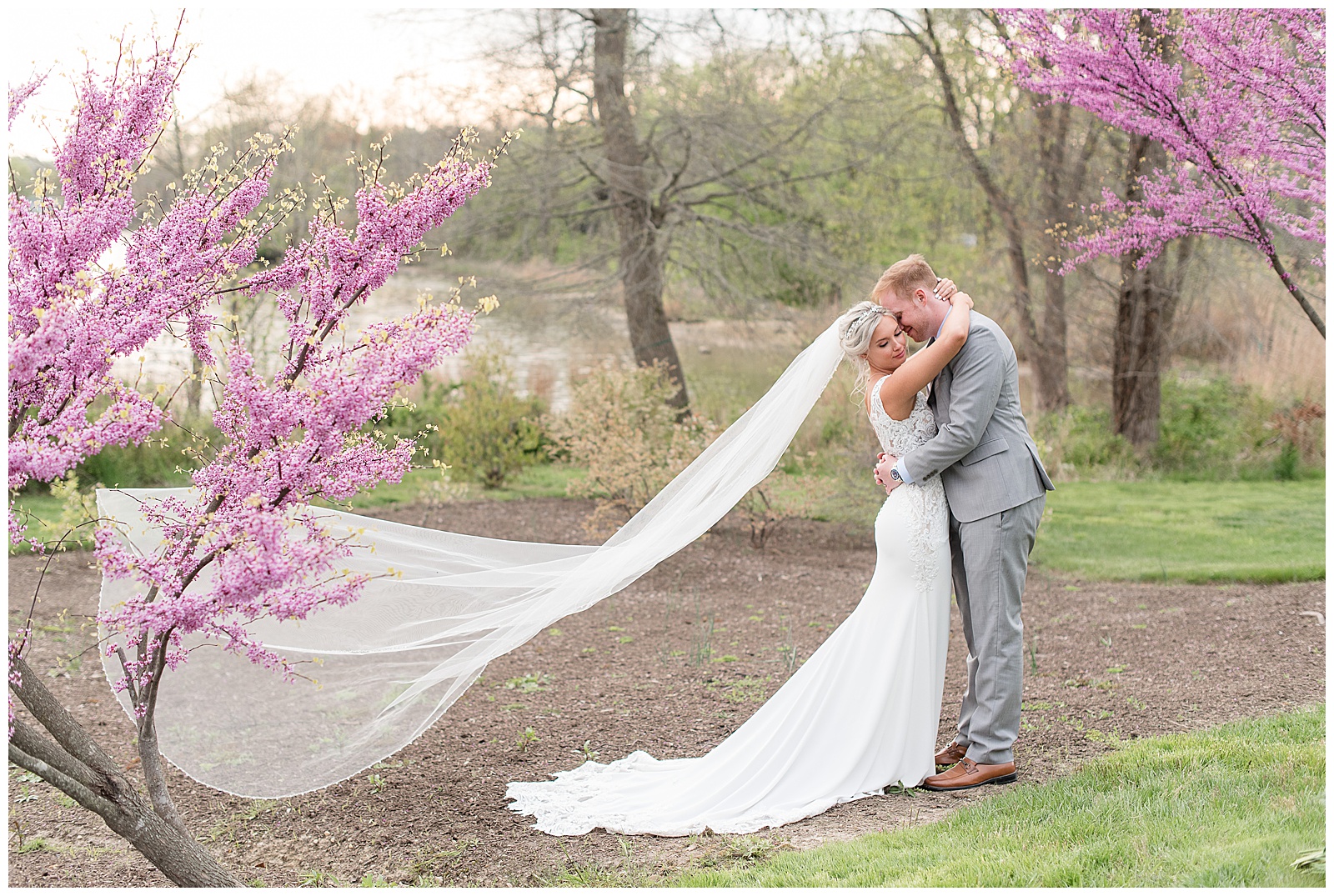 bride and groom hugging outdoors with long veil blowing behind her as they're surrounded by blooming eastern redbud trees