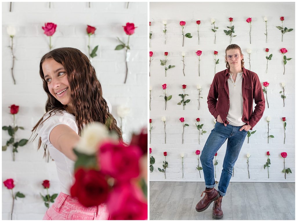 senior guy leaning against white wall with right knee bent in front of wall covered in red, pink, and white roses
