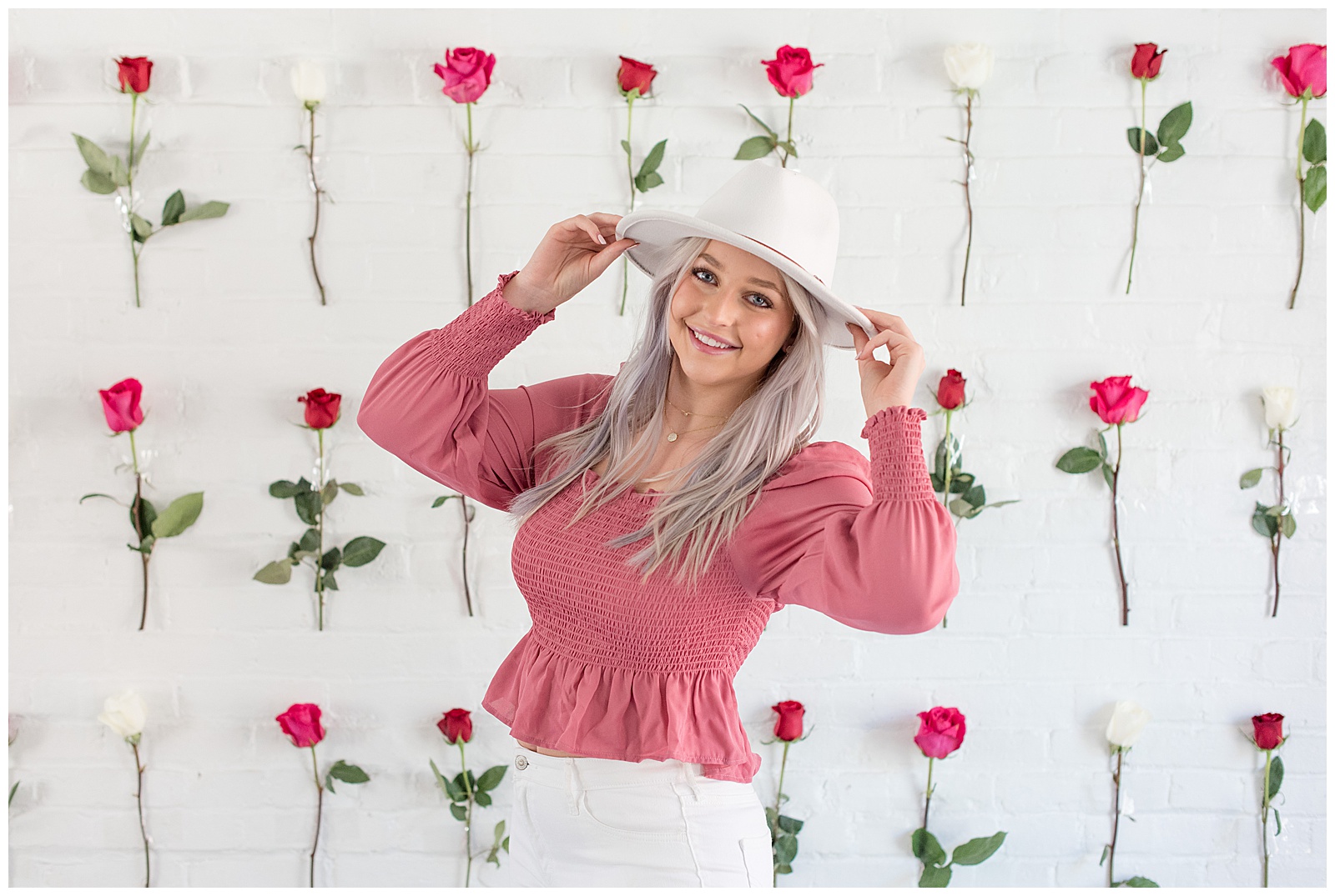 senior girl holding the brim of her white hat in front of white brick wall covered in roses at hingework
