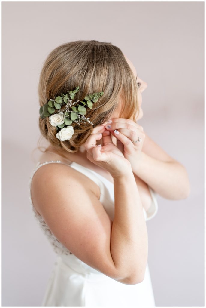 bride adjusting the back of her earring as she smiles in bridal suite in lititz pennsylvania with eucalyptus in her hair