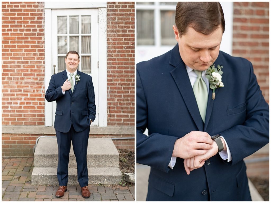 groom standing outside brick building by white door with his right hand holding onto edge of navy blue suit coat