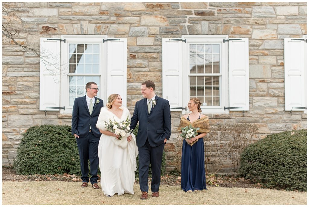 bride and groom holding hands and smiling with their best man and maid of honor beside them by stone building in lititz pennsylvania