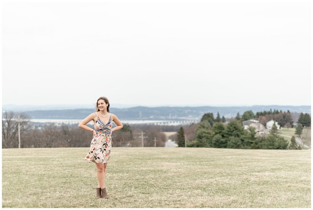 senior girl in floral dress with hands on hips looking right in large grass field in york pennsylvania