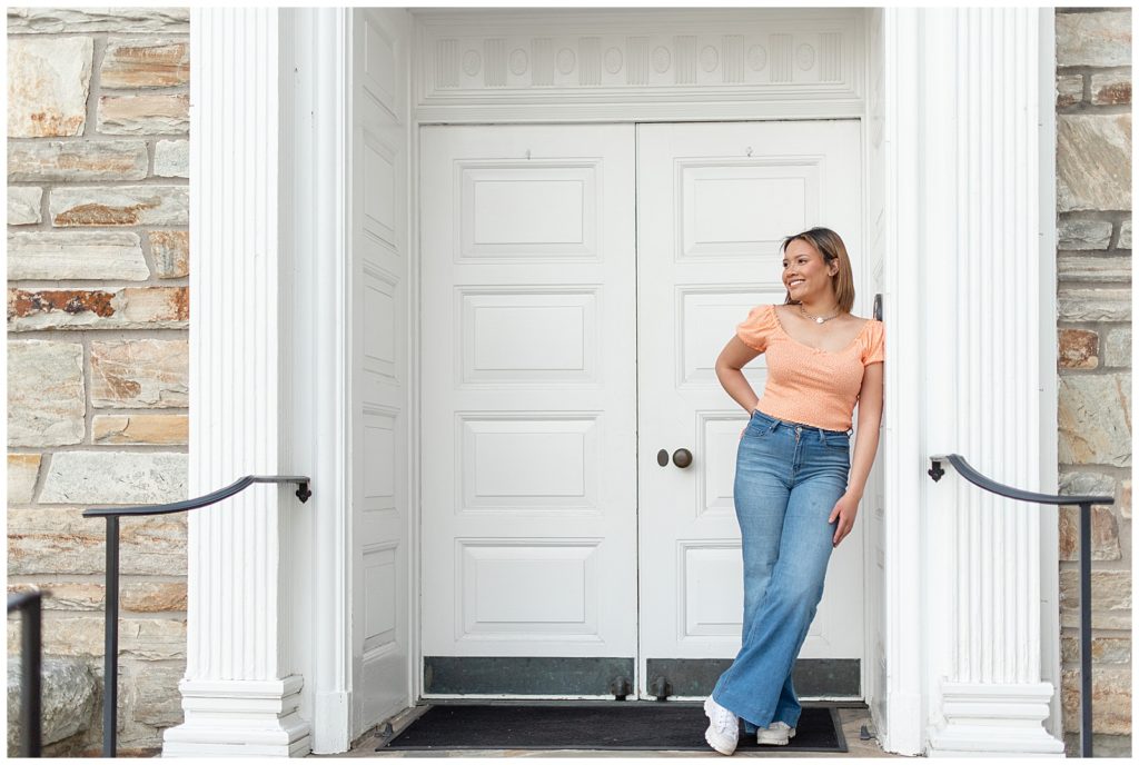senior girl in orange crop top and blue jeans leaning against white door frame in downtown lititz, pennsylvania