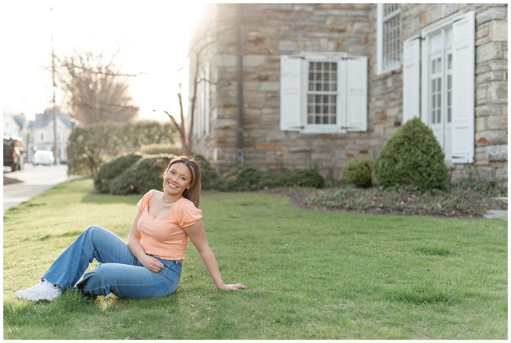 senior girl leaning back on her left hand while lounging in green grass by historic building in lancaster, pennsylvania
