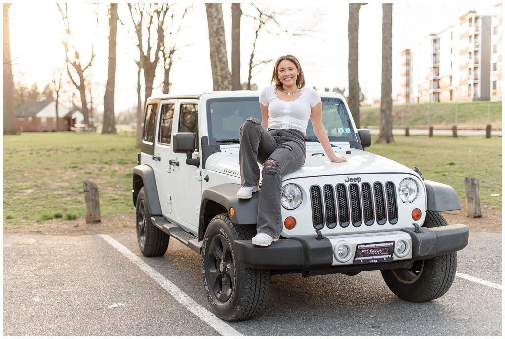 senior girl in white top and black jeans sitting on the hood of her white and black jeep wrangler at lititz springs park