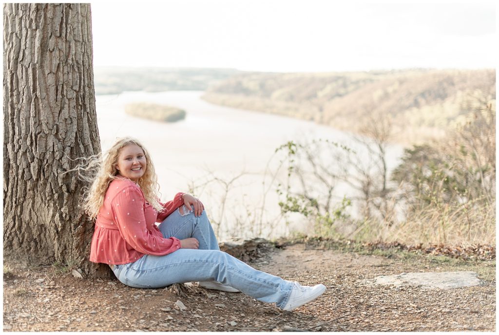 senior girl sitting on the ground leaning against tree atop pinnace point overlook by the susquehanna river below