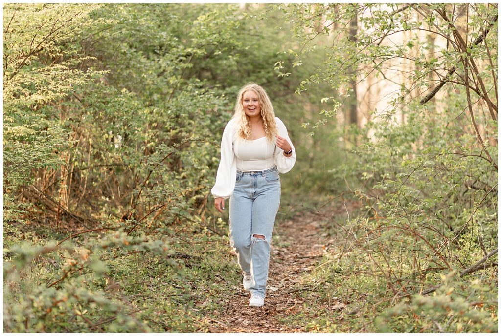 senior girl in white top and ripped blue jeans walking towards camera with left hand holding her long curly hair in woods in lancaster county