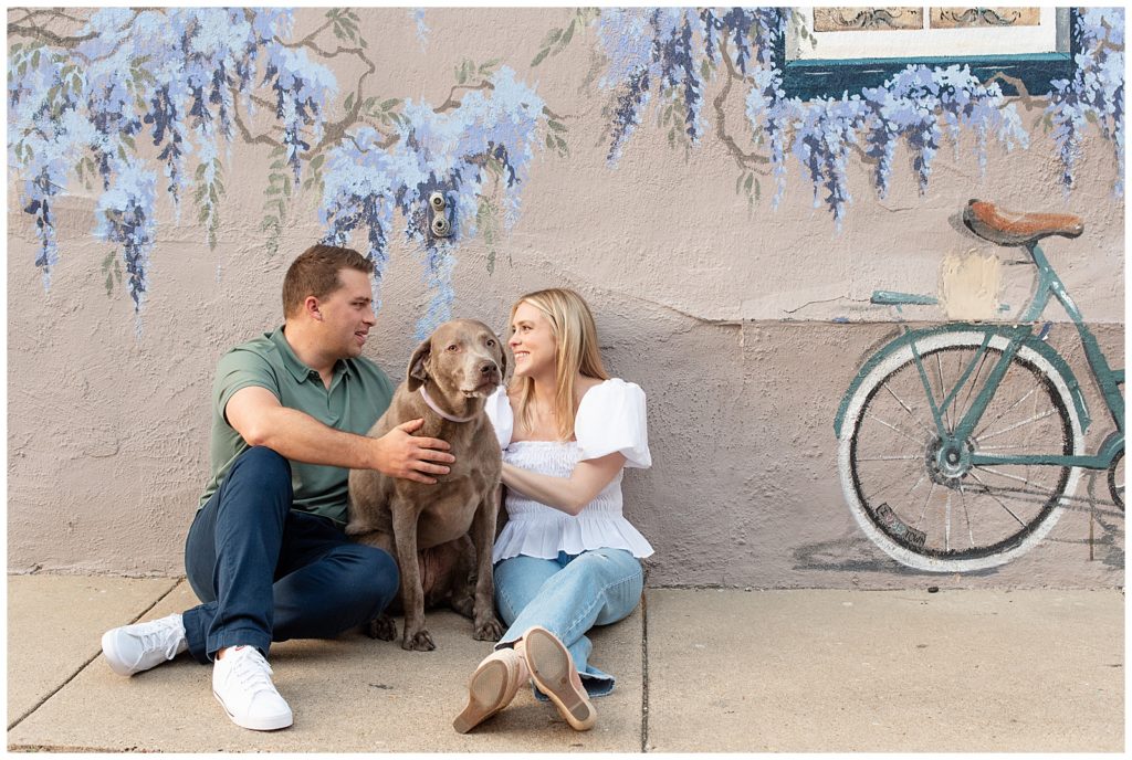 engaged couple sitting on sidewalk by painted wall with their dog between them in annapolis maryland