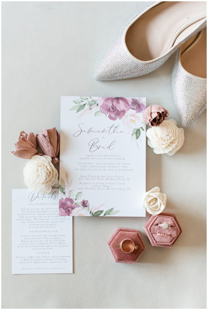 wedding invitation surrounded by mauve and white roses and wedding rings and bride's white high heels
