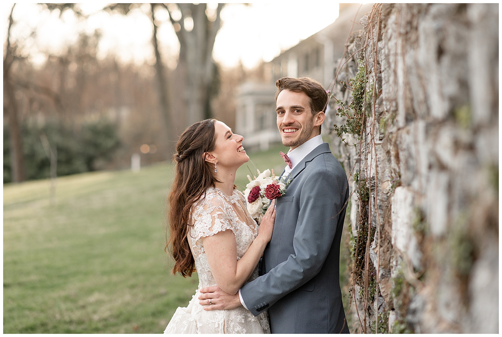 groom leaning back against stone wall and smiling at camera as bride looks and smiles at him in central pennsylvania