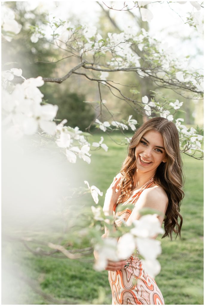 senior girl with left shoulder toward camera smiling as she stands behind blooming white tree at masonic village