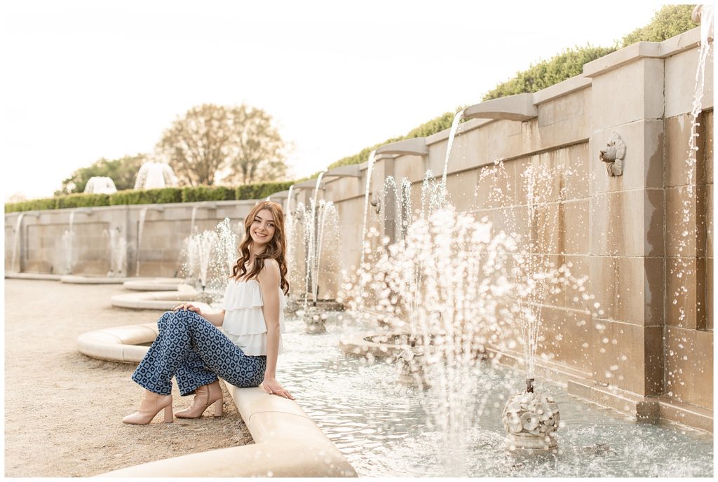 senior girl sitting on concrete step by beautiful fountain in white tank top and blue jeans at masonic village on spring evening