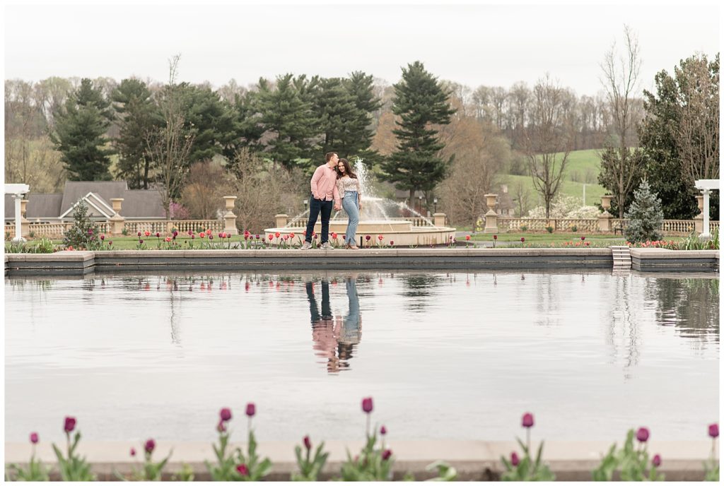 guy wearing pink shirt and dark jeans and girl wearing floral top and jeans kissing by pond at masonic village