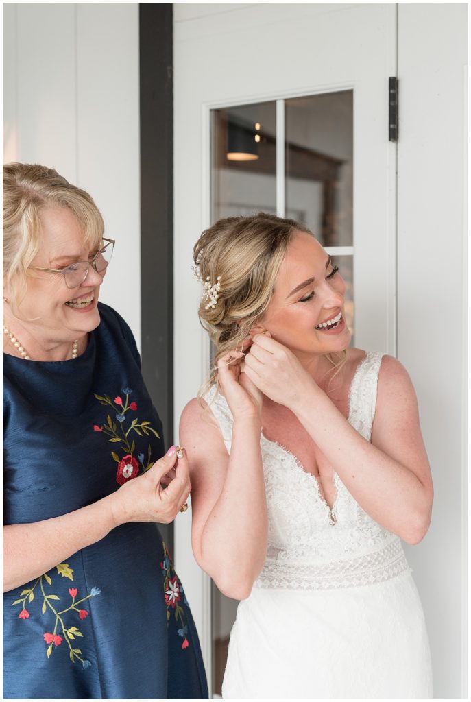 bride's mom and her sharing a laugh as bride puts earring into her ear in bridal suite at the barn at silverstone
