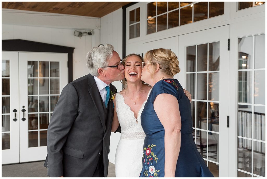 bride's parents on each side of her kissing her cheeks as she smiles at the barn at silverstone