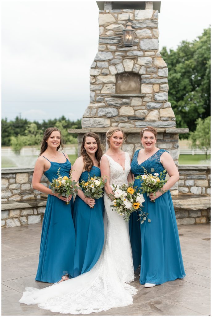 bride with her three bridesmaids wearing teal dress by outdoor stone fireplace at the barn at silverstone