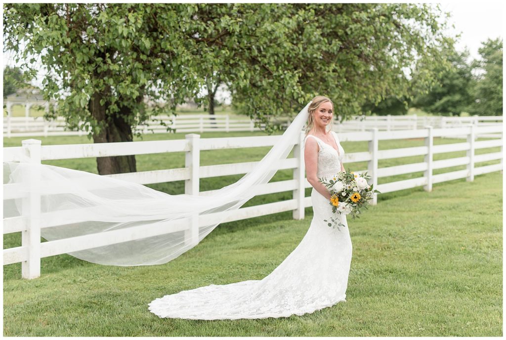 bride standing in lawn by white fence holding bouquet with long veil blowing behind her at barn venue in lancaster pa