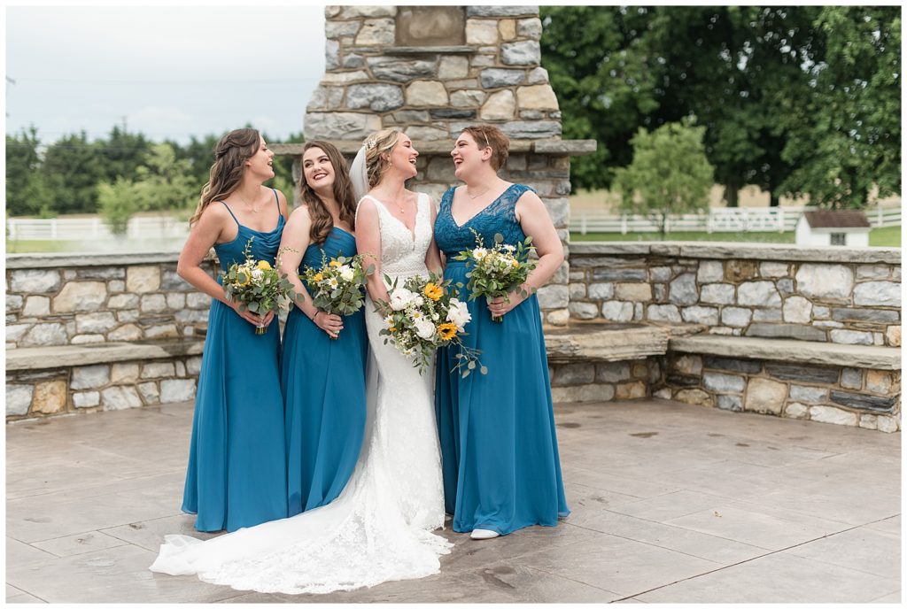 bride and her bridesmaids all smiling at looking at each other by outdoor stone fireplace at the barn at silverstone