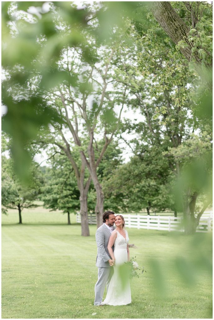 groom behind his bride outdoors by trees and white fence on sunny evening at the barn at silverstone