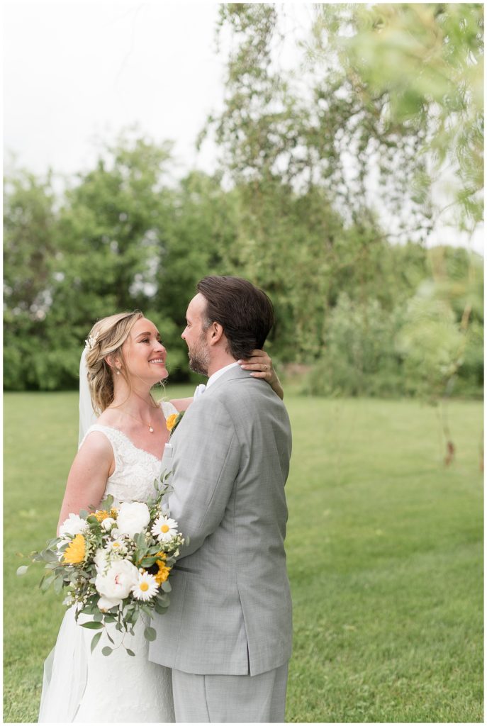 bride holding bouquet of white and yellow flowers as she hugs and stares at her husband outdoors at the barn at silverstone