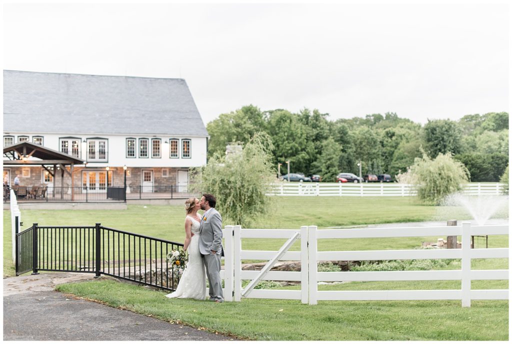 bride and groom kissing by fence with white barn behind them on sunny spring evening at the barn at silverstone