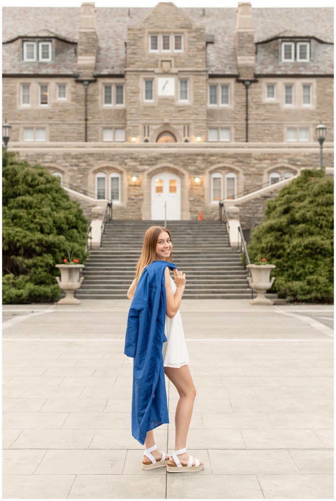 senior girl wearing white dress with her blue graduation gown draped over right shoulder in front of masonic village in lancaster county