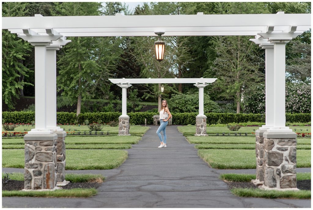 senior girl with white tank top and blue jeans standing under arbor with left knee popped at masonic village