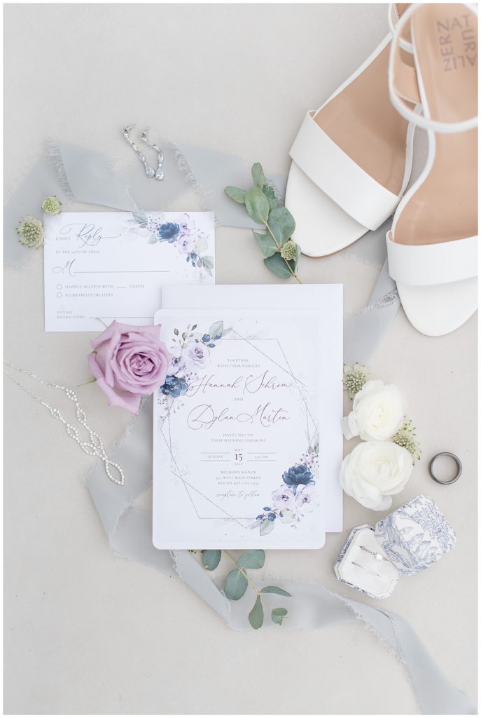 wedding invitation in sage green and purples surrounded by purple roses and eucalyptus and wedding rings and white sandals in lancaster county