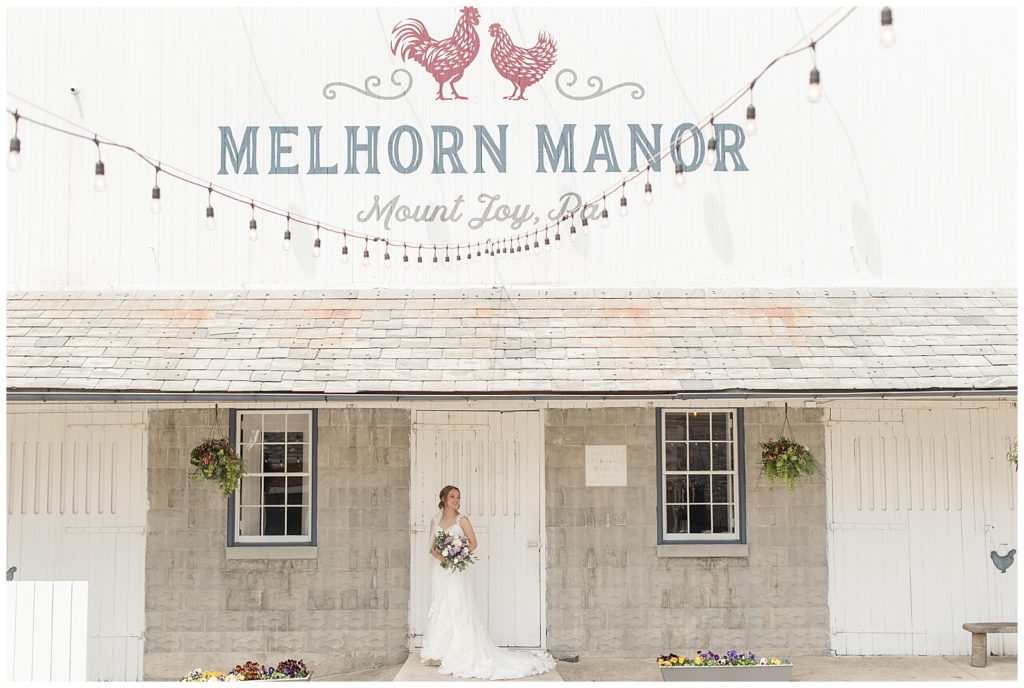 bride holding bouquet and standing by white barn door at melhorn manor barn on sunny spring day in mount joy pennsylvania