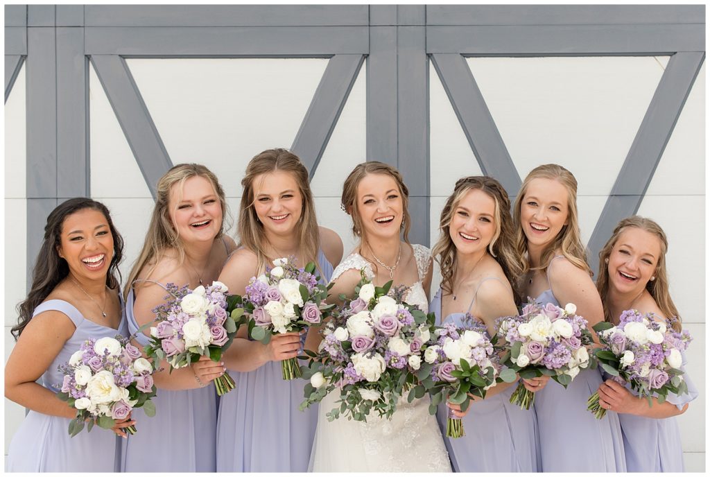 bride surrounded by her six bridesmaids all wearing light purple gowns and smiling big at melhorn manor